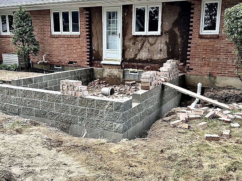 Concrete block foundation wall under construction against the exterior of a brick ranch house